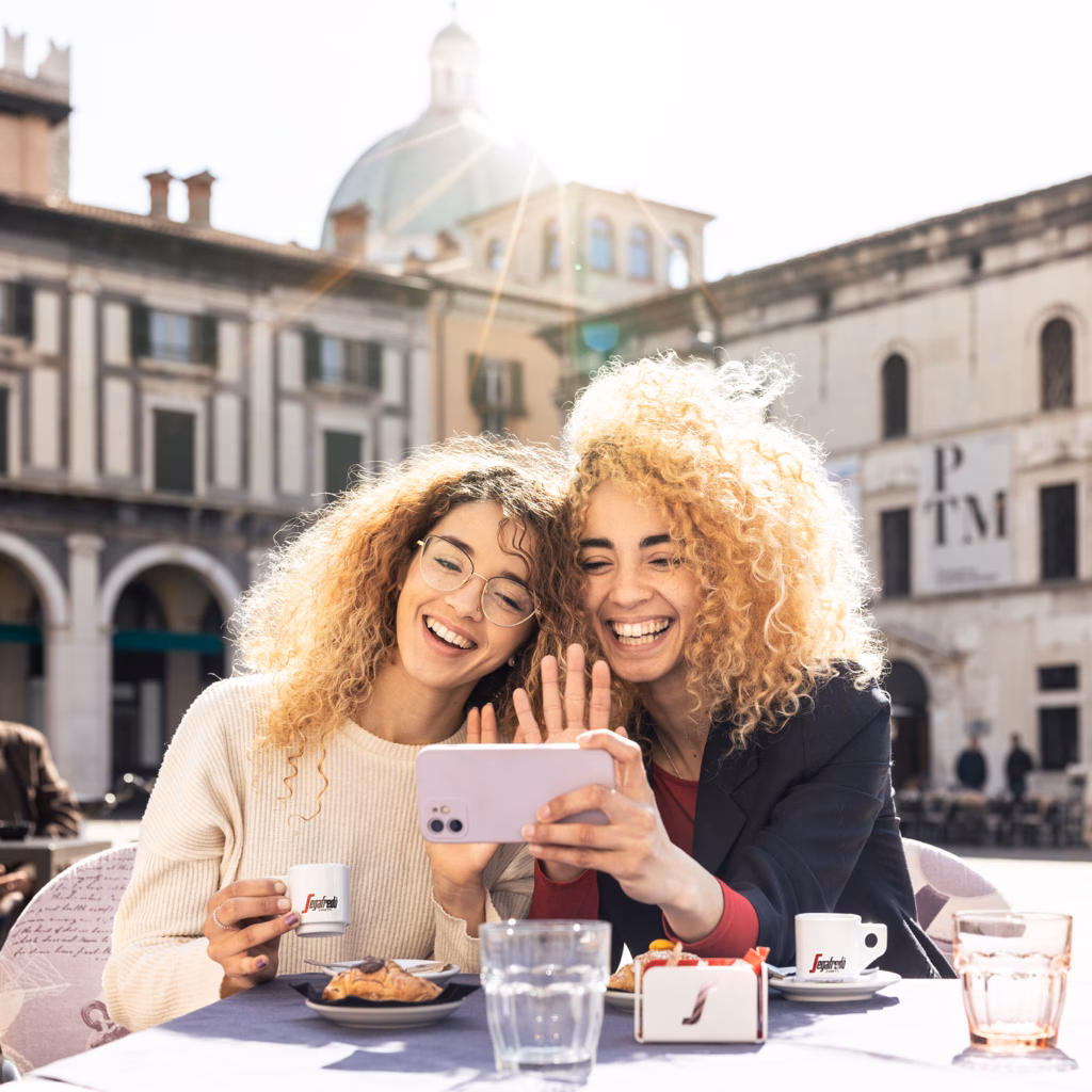 Imagebild: zwei fröhliche junge Frauen an einem Tisch auf einem Platz mit Kaffee, sie winken in ein Smartphone